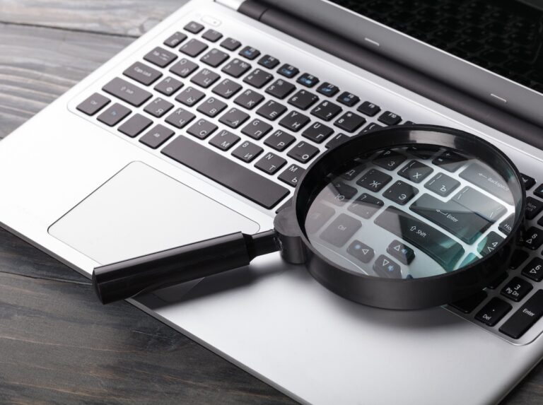 A looking glass is lying on a keyboard of a silver laptop computer with black keys. It is highlighting the Enter and Shift buttons.