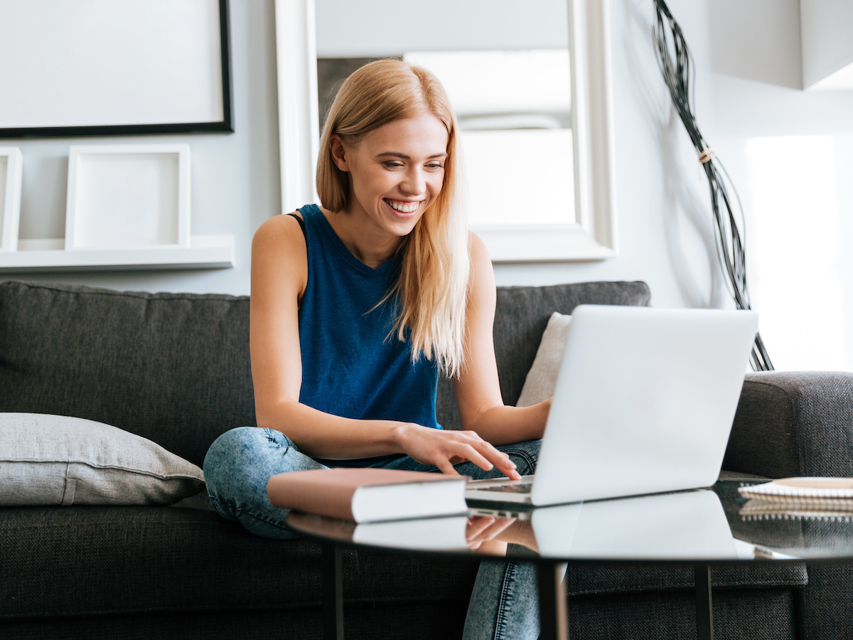 A strawberry blonde lady in her late 20's, wearing a navy blue tank top and blue jeans, is sitting casually on a grey couch. There is a silver laptop on a glass coffee table in front of her, as well as a beige book and a notebook. She is smiling at her laptop screen and touching the laptop's mousepad.
