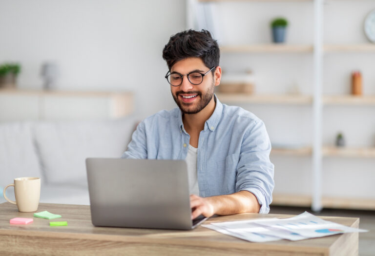 Smiling man using a laptop at a desk with papers and a coffee mug.