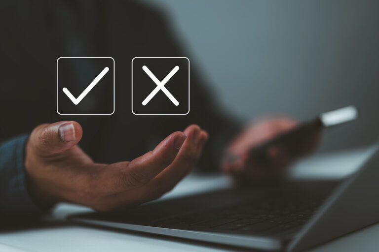 Business person holding a mobile phone with check mark and cross mark icons over a laptop.